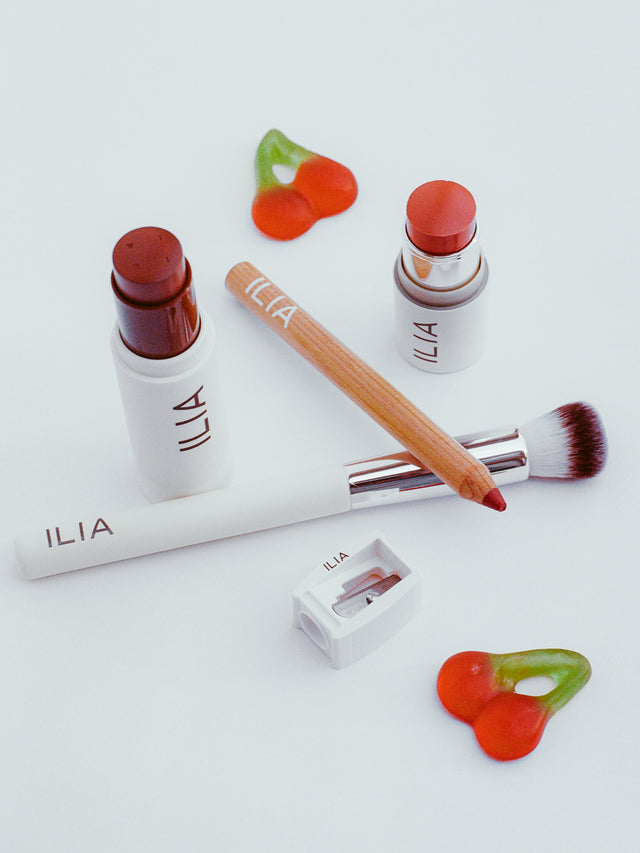 ILIA beauty products, including a brush, lip pencil, foundation stick, and a multi-stick, arranged with gummy cherry candies.